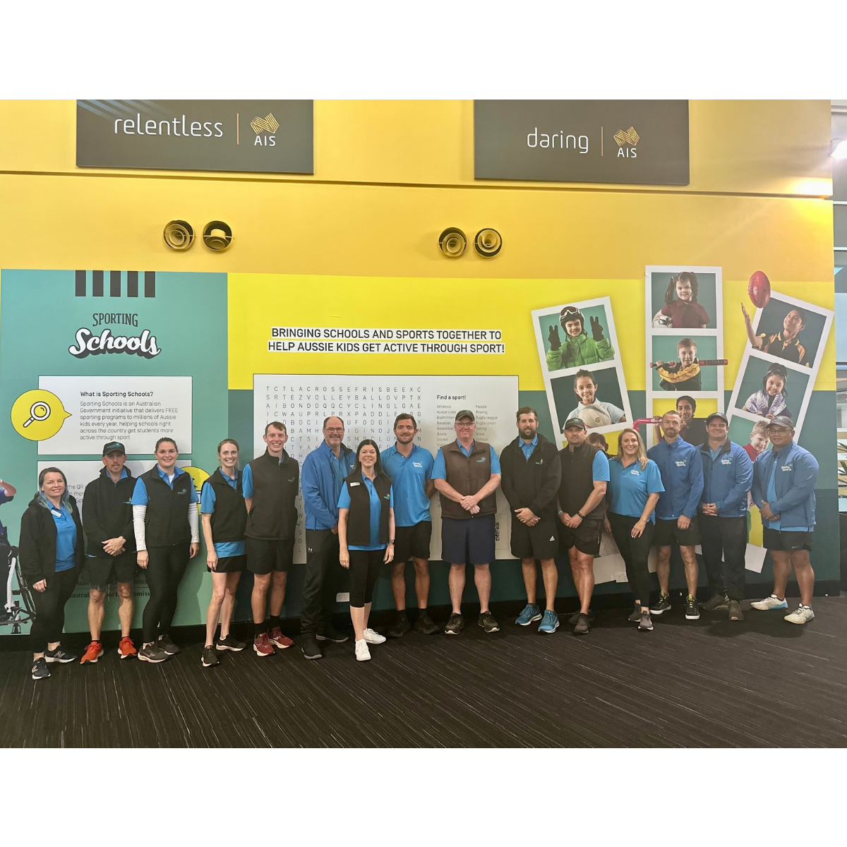The image shows a group of fifteen individuals posing together in front of a vibrant display with yellow and blue tones. The display is related to a program called "Sporting Schools," which aims to integrate sports into schools and encourage physical activity among Australian children. The group, wearing coordinated blue and grey sportswear, stands smiling before panels adorned with photos of children engaged in various sports, and informative texts like "Bringing schools and sports together to help Aussie kids get active through sport."