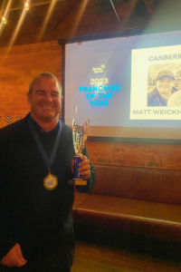 The image captures a man smiling proudly as he holds a trophy and wears a medal. He is standing in front of a screen displaying a presentation that reads "2023 Franchise of the Year" along with his name, "Matt Weickhardt," and the logo of Gecko Sports. The setting is a cozy venue with exposed brick walls and ambient lighting, enhancing the celebratory atmosphere of the event.