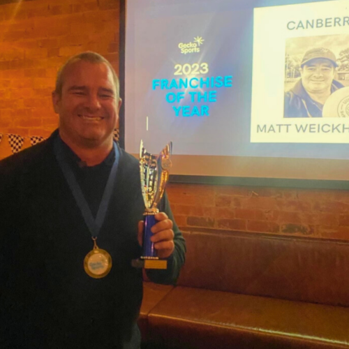 The image captures a man smiling proudly as he holds a trophy and wears a medal. He is standing in front of a screen displaying a presentation that reads "2023 Franchise of the Year" along with his name, "Matt Weickhardt," and the logo of Gecko Sports. The setting is a cozy venue with exposed brick walls and ambient lighting, enhancing the celebratory atmosphere of the event.