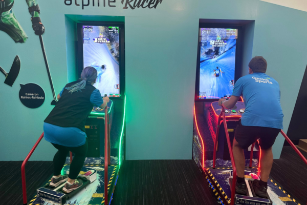 The image shows two people, a man and a woman, engaged in a virtual alpine skiing game. They stand on ski simulators with screens in front displaying fast-paced downhill races. The woman on the left is focused intently on her screen, leaning into a turn, while the man on the right, also concentrating, manoeuvres through the virtual course. The area is decorated with ski-themed elements, and the names of athletes, such as "Cameron Rahles-Rahbula," are displayed, adding a personalised touch to the sports training environment.