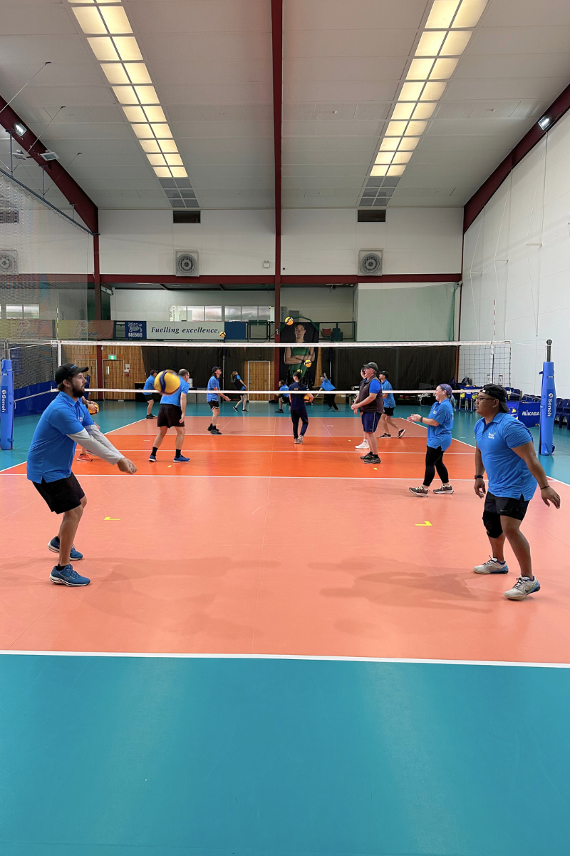 The image features a vibrant indoor volleyball game with eight players, all dressed in blue shirts and various shades of dark pants or shorts. They are actively engaged in the game, with some players ready to hit the ball and others in motion, positioned on an orange court. The high ceiling of the facility is equipped with bright lights and a banner that reads "Fuelling excellence," suggesting a focus on sports training or competition excellence. The atmosphere is lively and energetic, indicative of a team sport event.