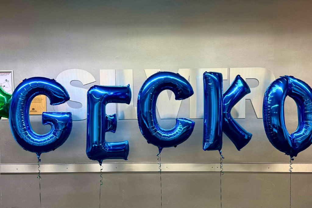The image features shiny blue metallic balloons spelling out "GECKO" against a neutral grey wall. The letters are bold and inflated, creating a festive and celebratory atmosphere. The reflection on the balloons adds to their glossy appearance, enhancing the visual impact of the setup.