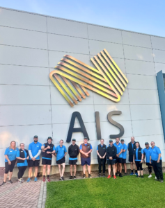 The image shows a group of fourteen people posing in front of a large building with a prominent "AIS" logo. The logo, depicted in stylised golden strips, adds a dynamic element to the simple, modern architecture of the building. The individuals, dressed in coordinated blue team apparel, display a mix of smiles and relaxed postures, indicating a sense of camaraderie or team spirit.
