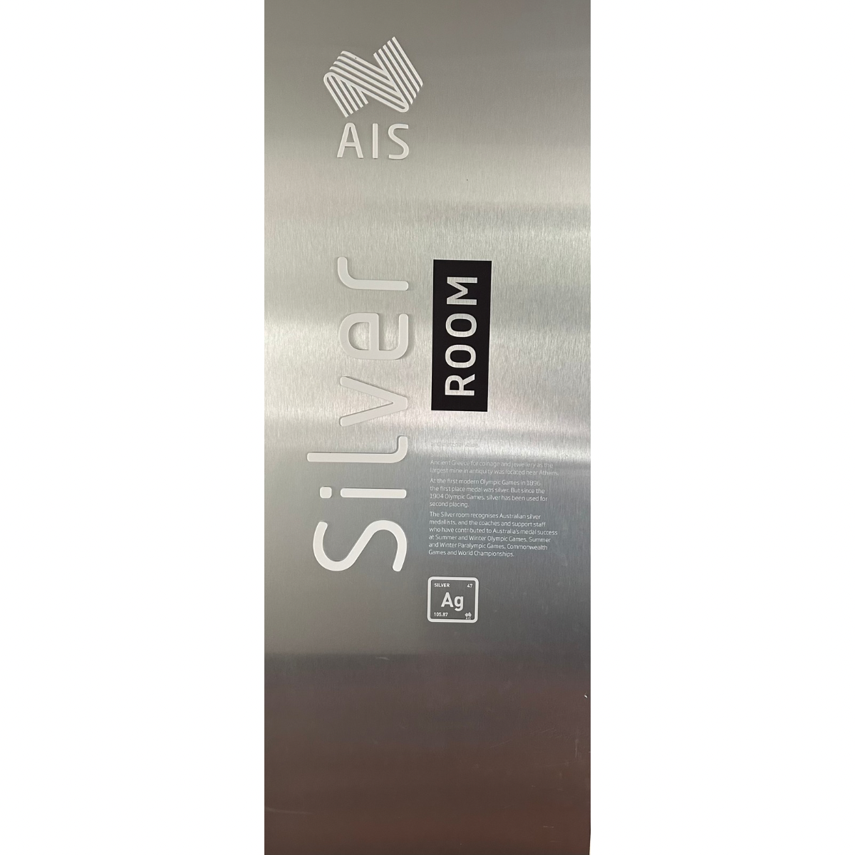 The image shows a sleek, metallic door sign reading "Silver Room" with a large "AIS" logo above it. Below the room name, there's a detailed description explaining the significance of silver in relation to the room, presumably referring to its attributes or the theme it represents. To the lower right corner of the sign, there is a chemical symbol for silver, "Ag," adding a scientific element to the aesthetic.
