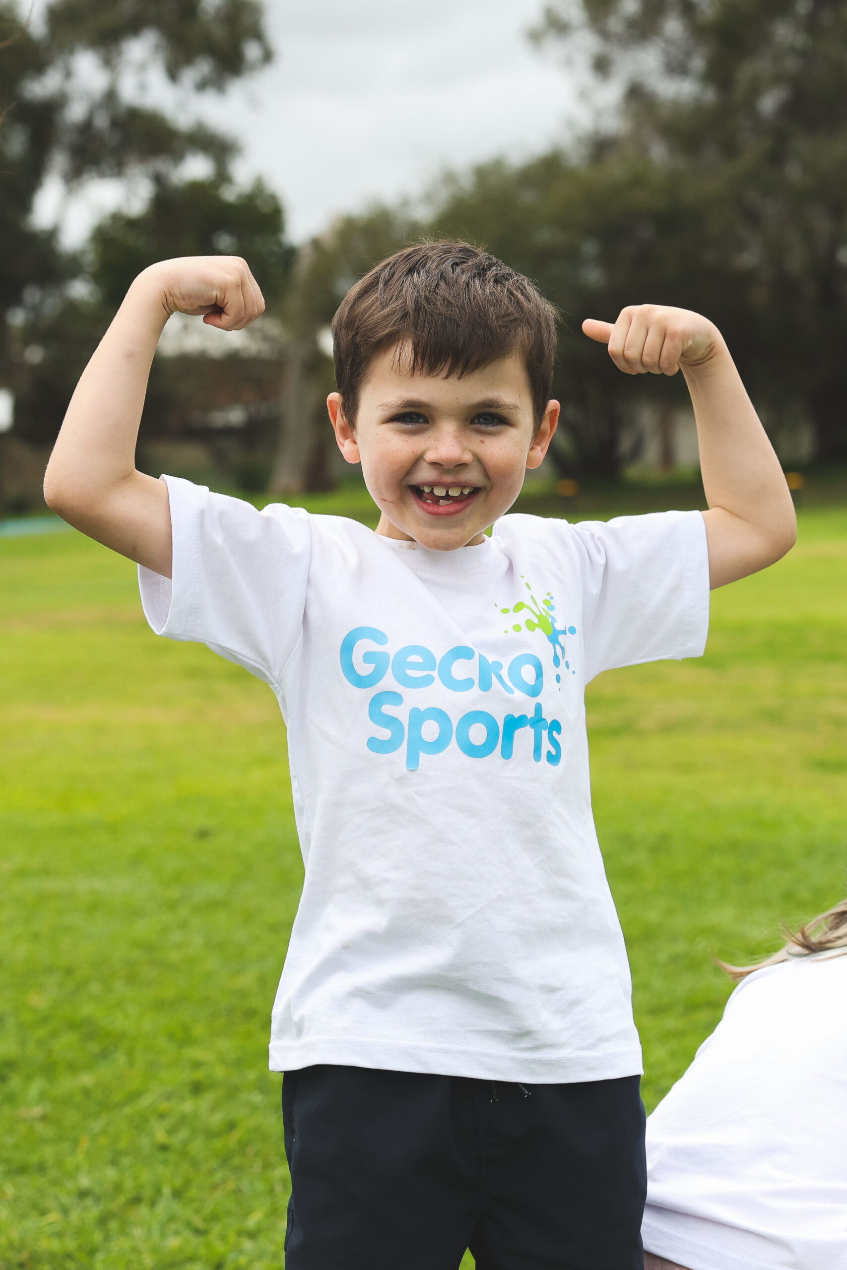 Child in a GeckoSports t-shirt smiling and flexing their arms on a grassy field.