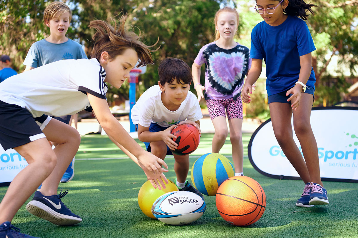 Children playing a multi-sport ball game on a turf field with several sports balls on the ground.