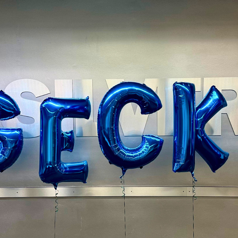 The image features shiny blue metallic balloons spelling out "GECKO" against a neutral grey wall. The letters are bold and inflated, creating a festive and celebratory atmosphere. The reflection on the balloons adds to their glossy appearance, enhancing the visual impact of the setup.
