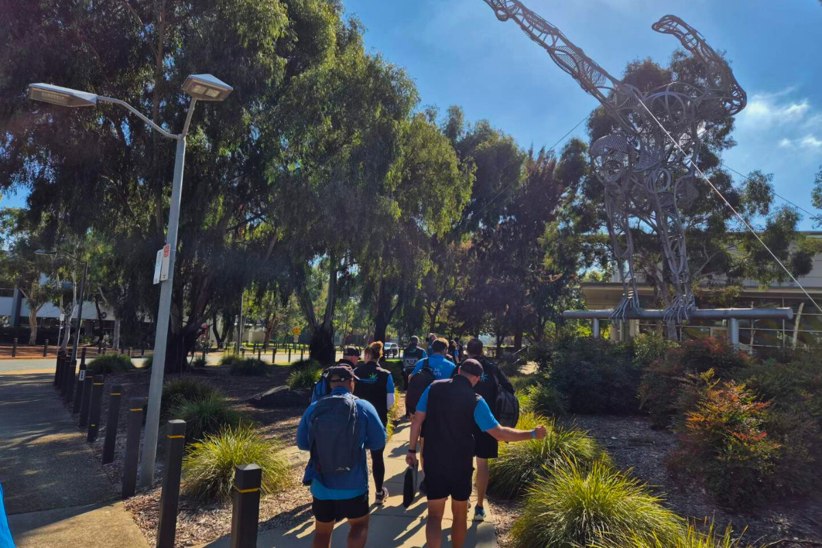The image depicts a group of people, mostly in blue shirts, walking along a path lined with lush greenery and modern street lamps. They are approaching or passing by a striking, large-scale sculpture that resembles a humanoid figure made from intertwined metallic parts, positioned prominently in the landscape.