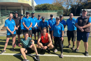 The image features a group of twelve individuals posing together on a sunny day at a sports facility. Most of them are dressed in matching blue Gecko Sports shirts, suggesting they are part of a team or staff. Three individuals in the front row are crouching, with the rest standing behind them, all smiling towards the camera, indicating a cheerful, team-oriented environment. The background includes a green fence and a part of a stadium, enhancing the sports theme of the photograph.